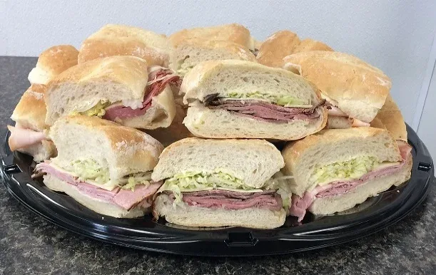 A black platter piled high with halved sandwiches on soft white buns, filled with sliced deli meat, cheese and shredded lettuce