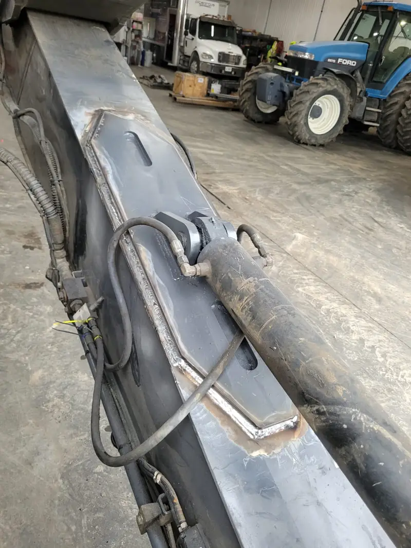 Excavator boom on the shop floor, prepared for welding, with tractors parked behind