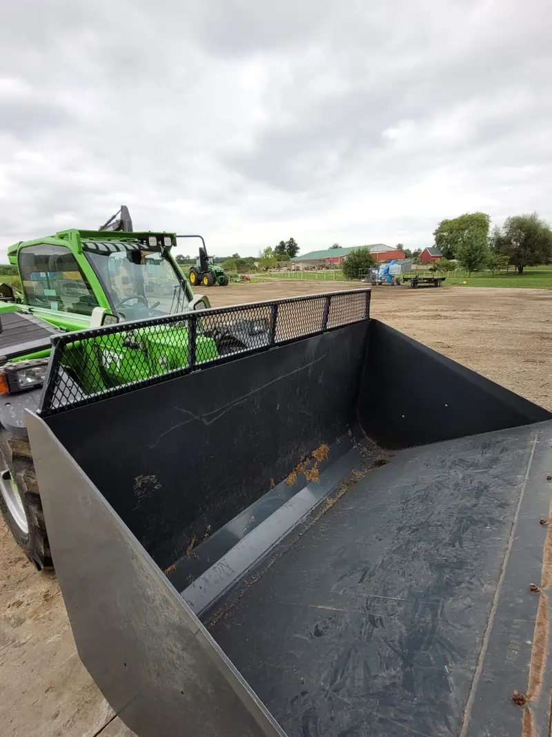 Large bucket with a mesh spill guard, mounted on a green telehandler in a gravel yard