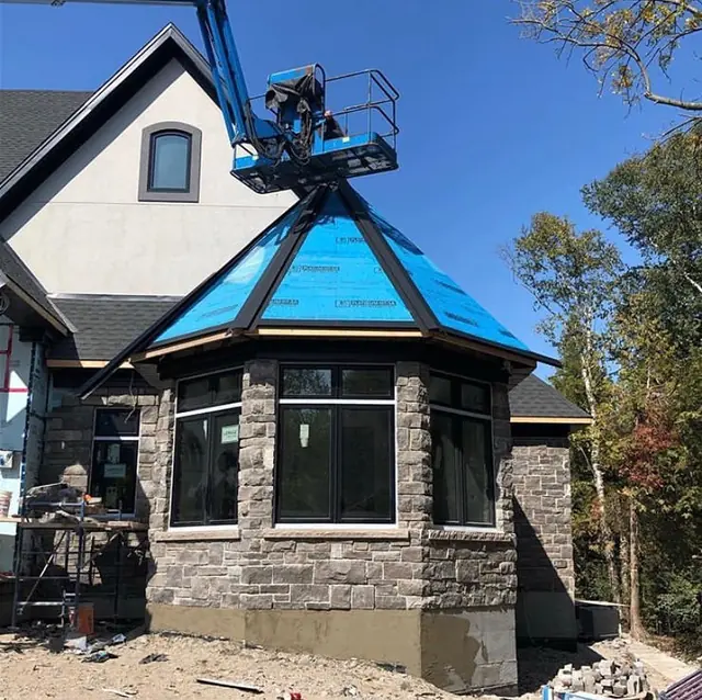 A house turret wrapped in bright blue roofing underlayment with a boom lift beside it, stone walls and new windows below