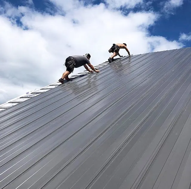 Two people working on a long grey standing seam steel roof under a blue sky with white clouds