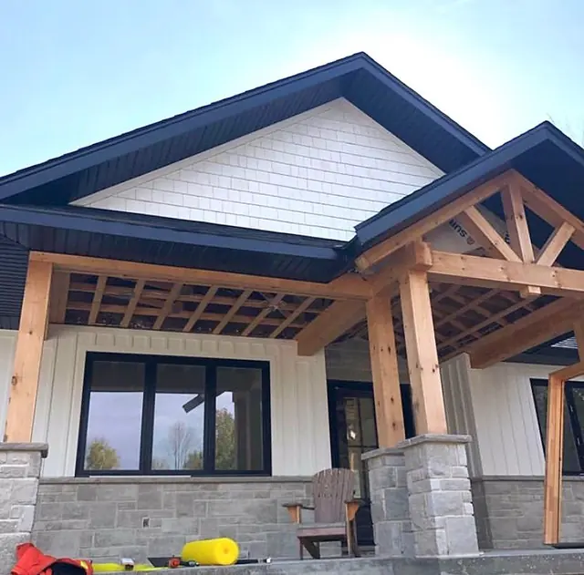 A house front with white shakes in the gable, white board and batten walls, a timber-frame porch with stone pillars and dark trim