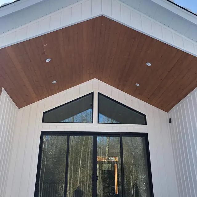 Looking up at a white board and batten gable with a stained wood ceiling, tall gable windows and black-framed patio doors
