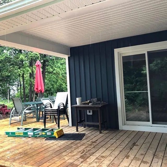A porch with white soffit and a white-clad beam over a wooden deck, navy vertical siding and a ladder lying on the deck