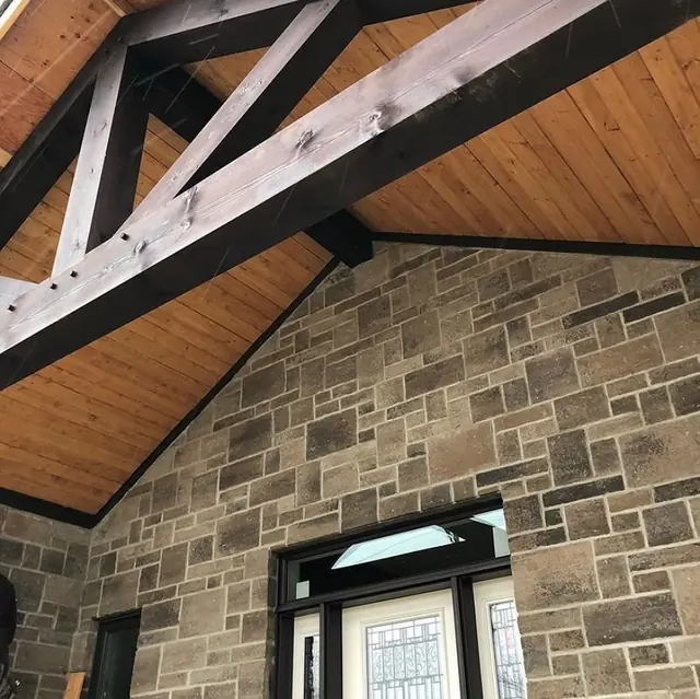A tongue-and-groove wood porch ceiling with dark stained truss beams above a stone wall and front door