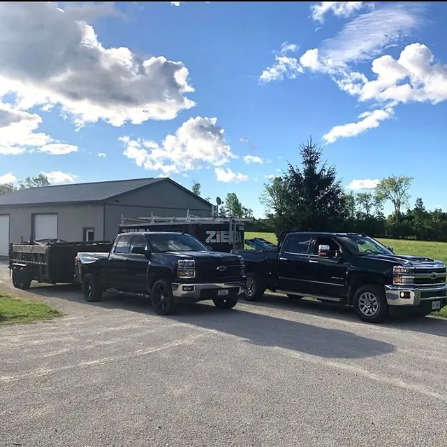 Two black pickup trucks, a dump trailer and a lettered work trailer parked on gravel beside a grey steel building