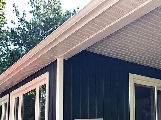 A white seamless gutter running along a cottage eave above navy vertical siding and white-trimmed windows, pine trees behind